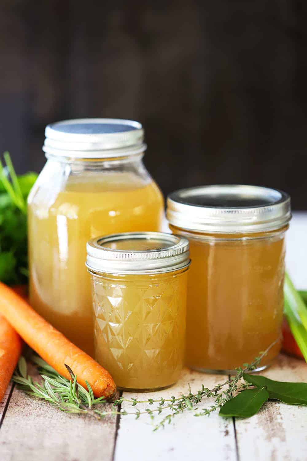 Three jars, ranging in size from small to large, filled with homemade chicken broth with loose carrots and herbs nearby. 