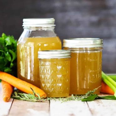 Three jars, ranging in size from small to large, filled with homemade chicken broth with loose carrots and herbs nearby.