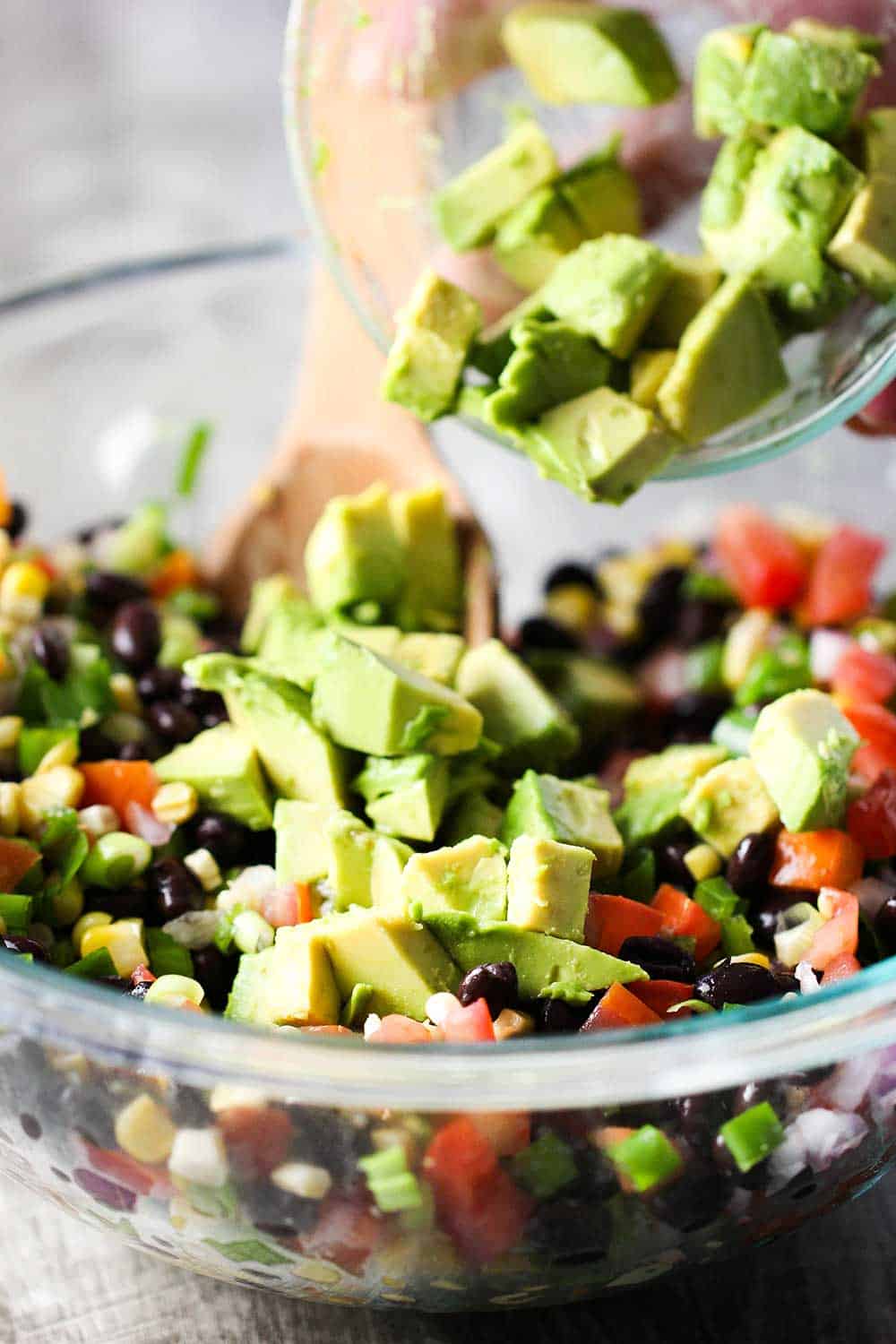 Fresh produce, including chopped avocado is mixed together in a large glass bowl. 