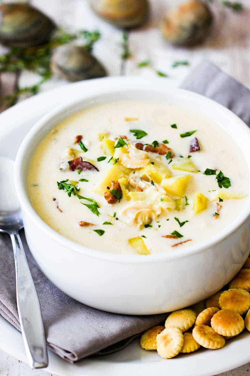 A large white soup bowl filled with New England Clam Chowder on a plate with oyster crackers and spoon next to it. 