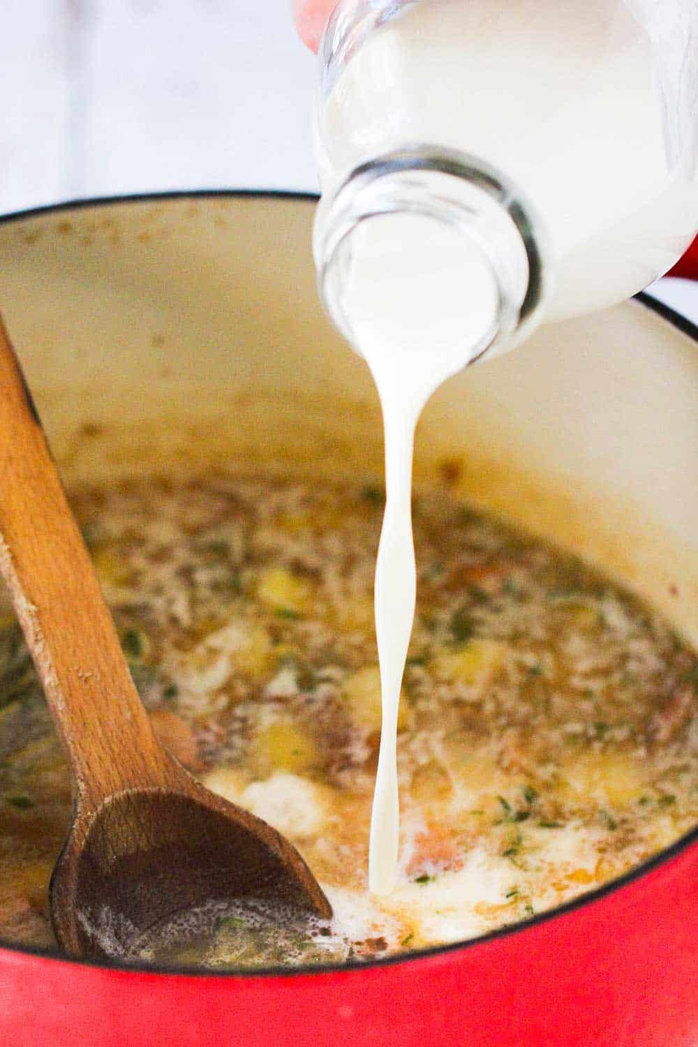 A hand pouring cream from a milk bottle in a Dutch oven filled with New England Clam Chowder. 