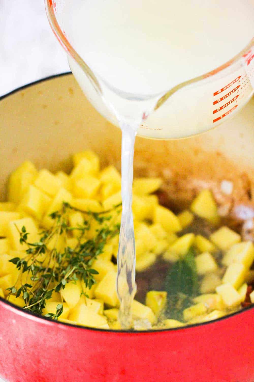 A hand pouring clam broth into a red Dutch oven filled with potatoes and herbs. 