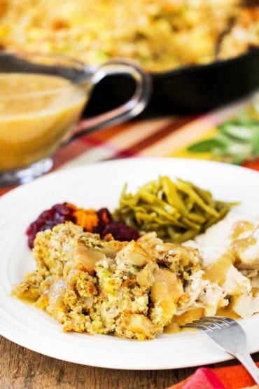 A white plate of Thanksgiving dressing, turkey, green beans, next to gravy and a black skillet.