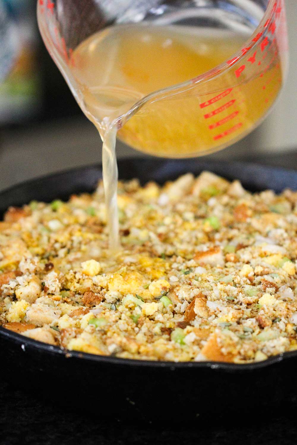 A hand pouring a large measuring cup of turkey stock into a pan filled with dressing. 