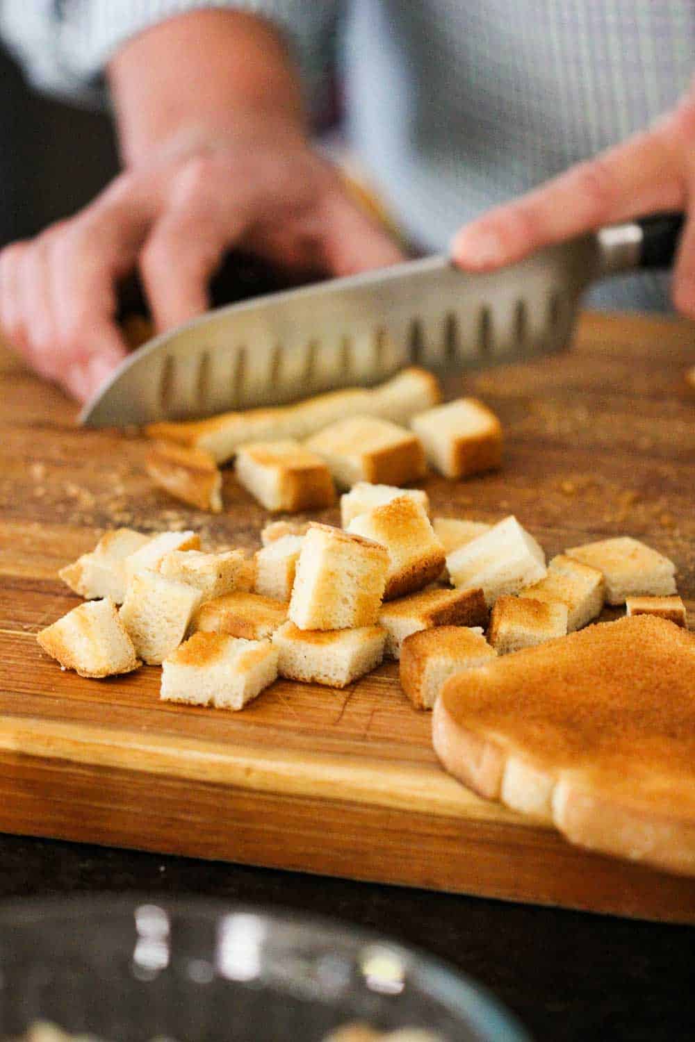 A hand using a knife to cube toasted white bread on a cutting board. 