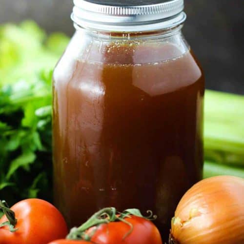 Homemade beef stock in a jar surrounded by tomatoes, celery, and other vegetables.