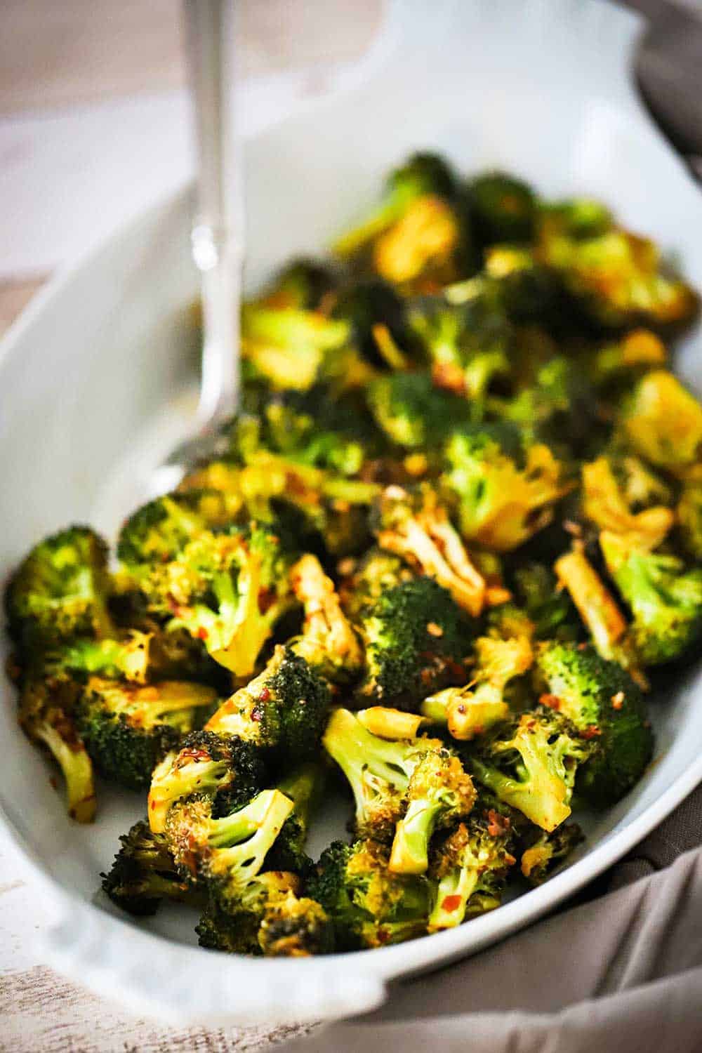 Roasted broccoli with red pepper flakes in an oval white serving platter with a silver spoon inserted into the dish.