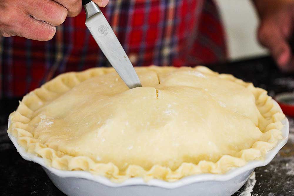 A hand using a knife to cut slits in the top of an uncooked homemade apple pie. 