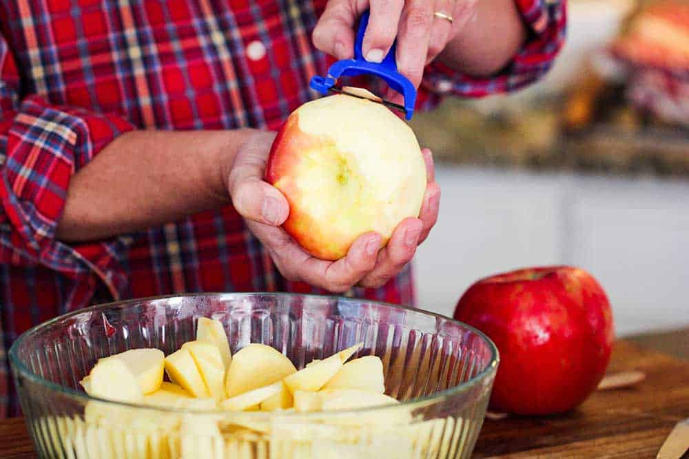 Two hands peeling a honeycrisp apple over a large bowl of slice apples. 
