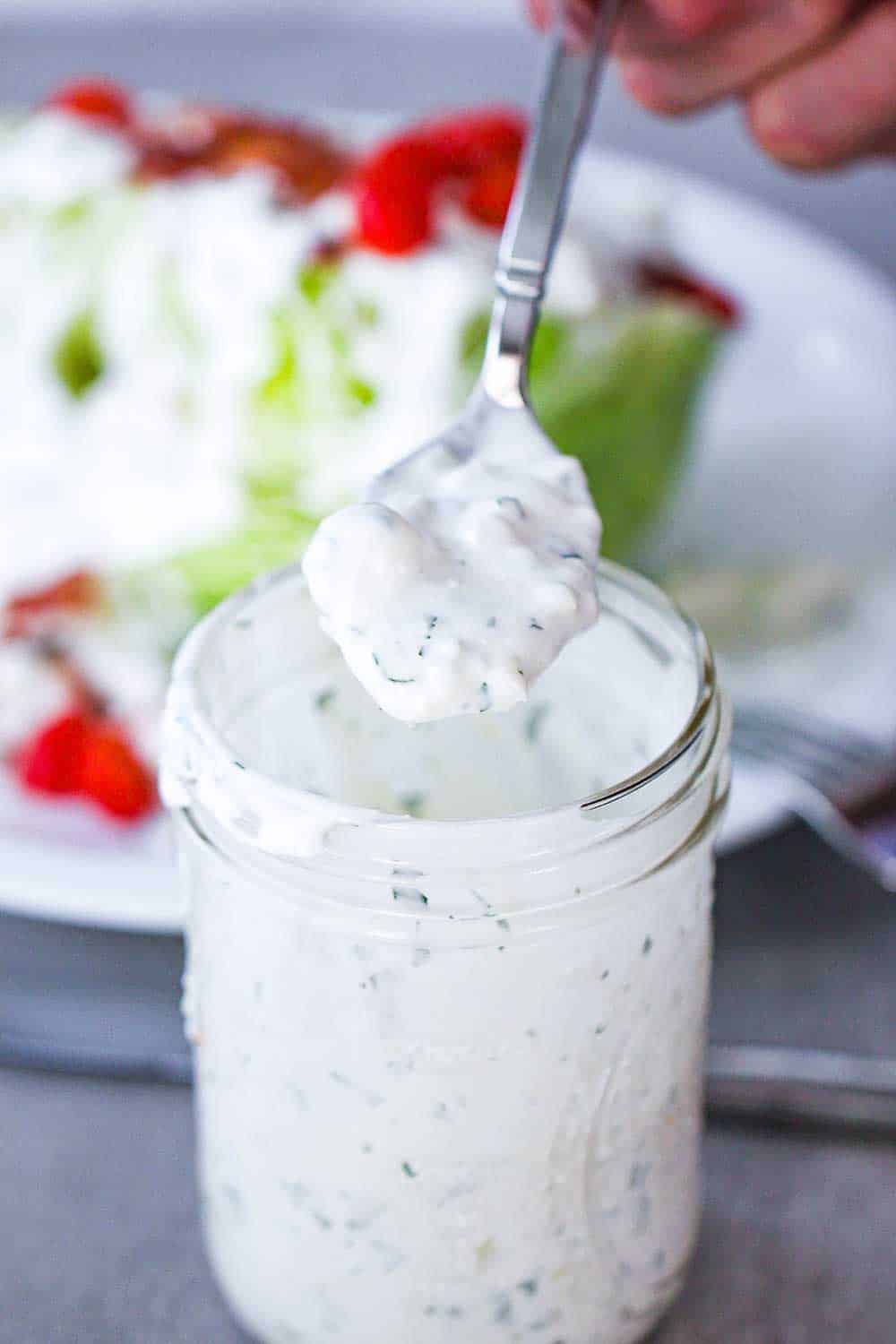 A spoon of blue cheese dressing over a jar with a salad in the background. 