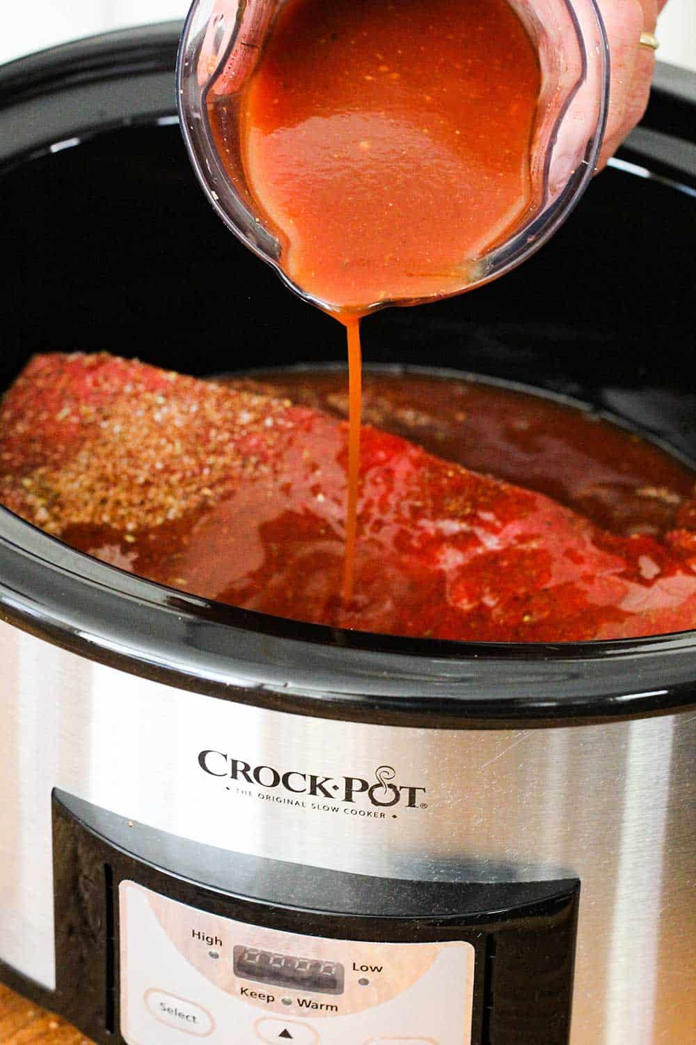 Homemade BBQ sauce being pour over a brisket in a Crock-Pot slow-cooker for BBQ brisket sandwiches.