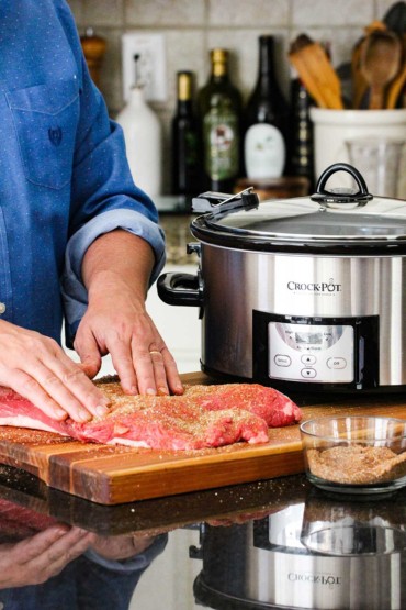 Hands adding a BBQ rub to brisket next to a Crock-Pot slow cooker.