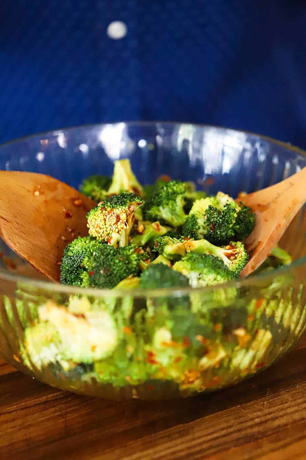 Two hands using two wooden spoons to toss broccoli florets in a spicy sauce in a large glass bowl. 