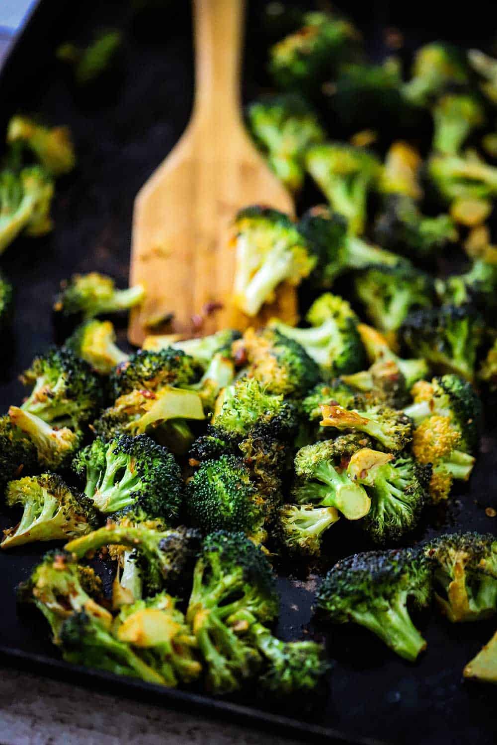 A baking sheet filled with roasted broccoli with a wooden spatula in the middle of the pan. 