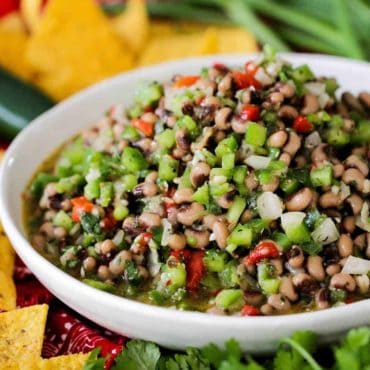 A large white bowl filled with Texas caviar surrounded by tortilla chips and scallions.