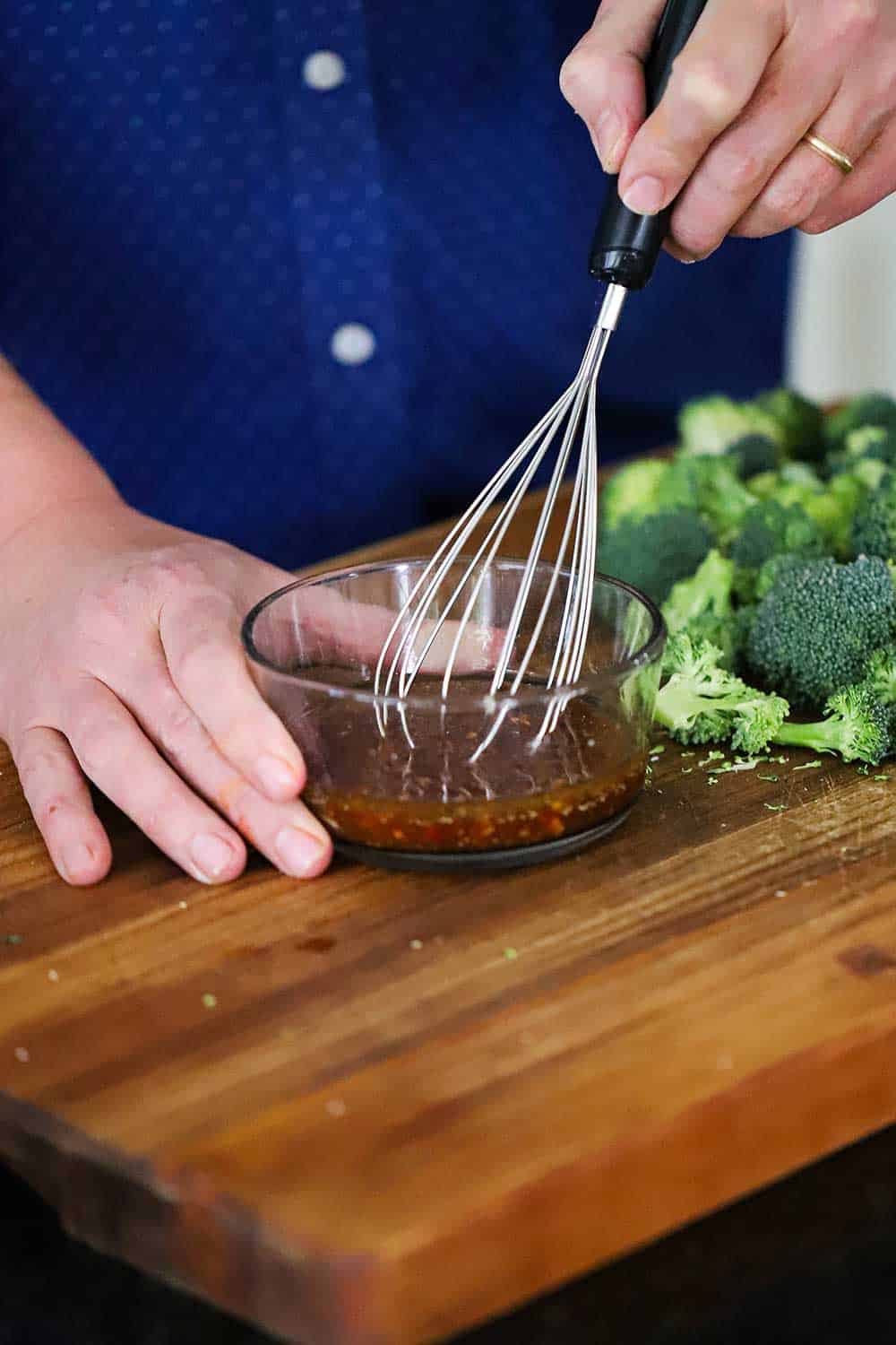 A person using a wire whisk to mix an chili paste and soy sauce mixture in a small glass bowl next to broccoli florets. 