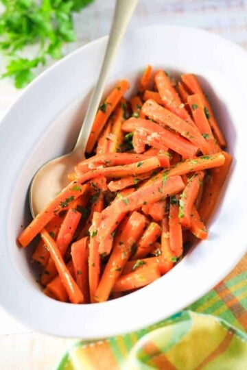 A white serving dish filled with maple braised carrots with a spoon in the bowl next to a sprig of parsley.