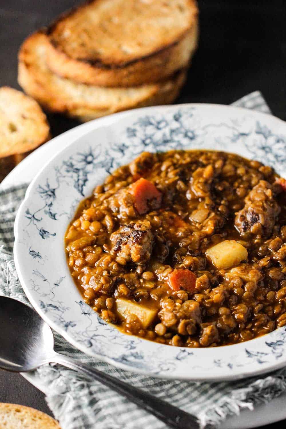 A white bowl of rustic lentil soup with slices of toasted bread stacked next to it.