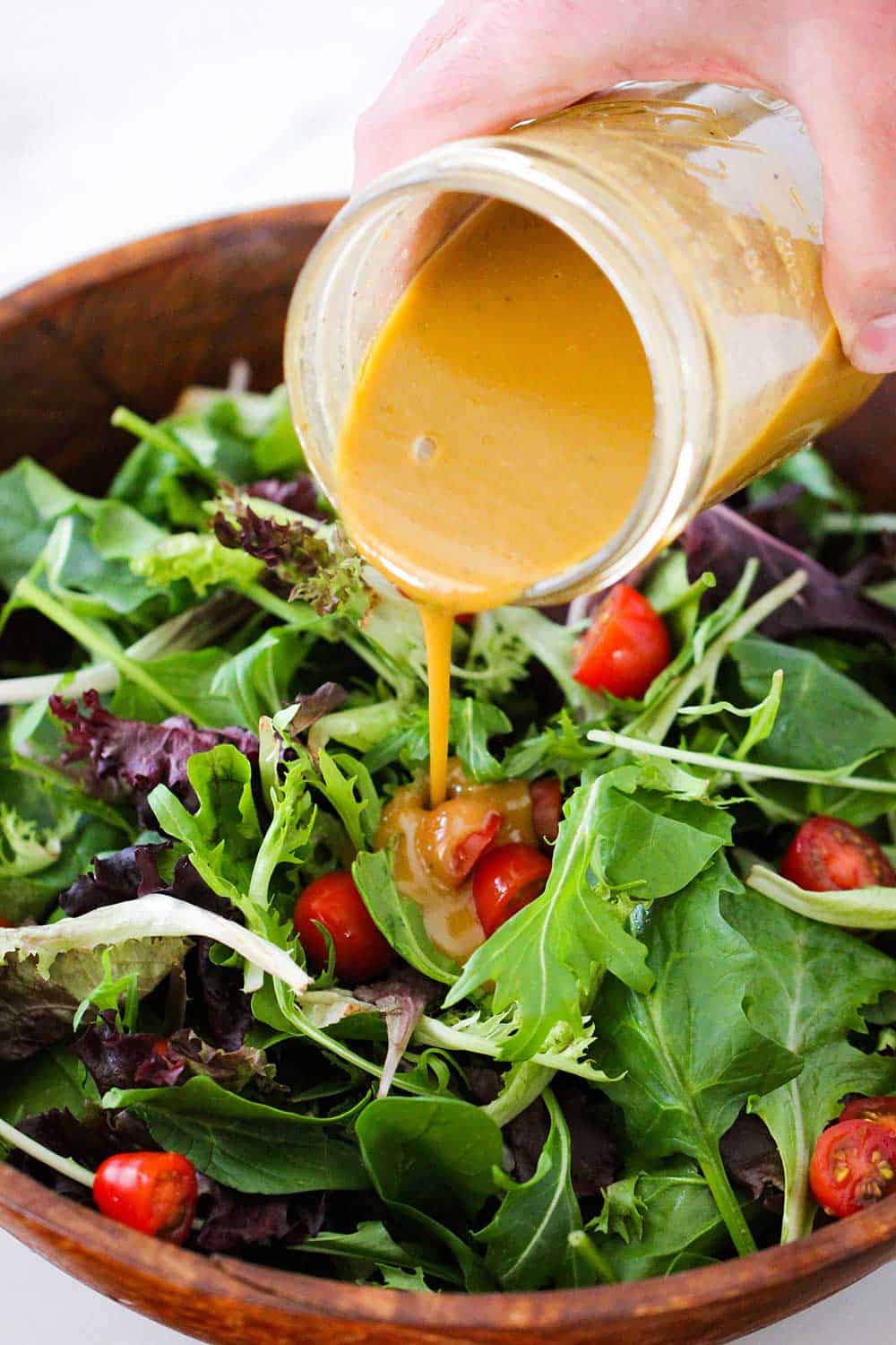 A hand pouring homemade balsamic vinaigrette from a jar onto a bowl filled with a green leaf salad. 