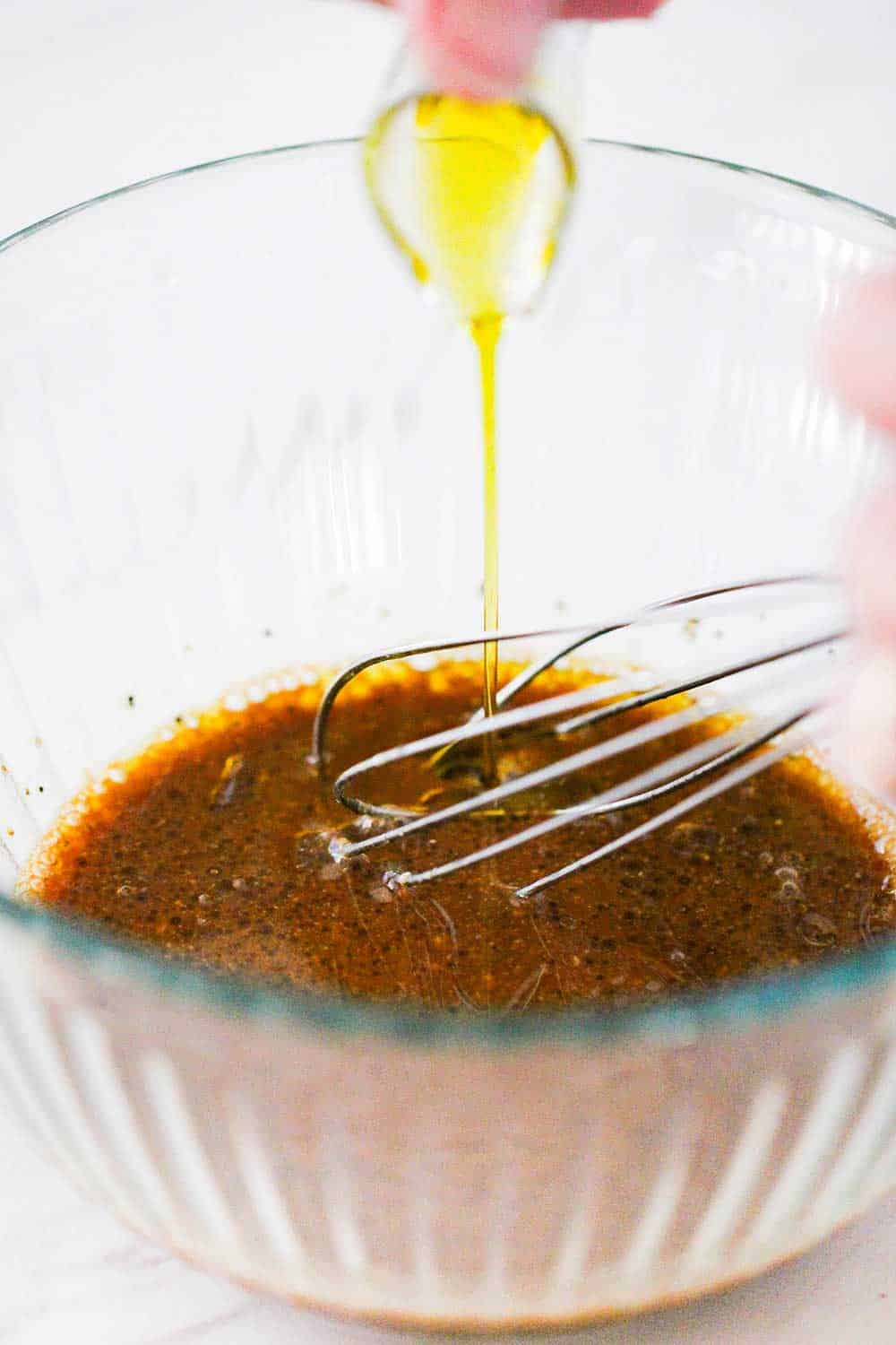 A whisk in a bowl of balsamic vinegar and other ingredients with oil being poured in. 