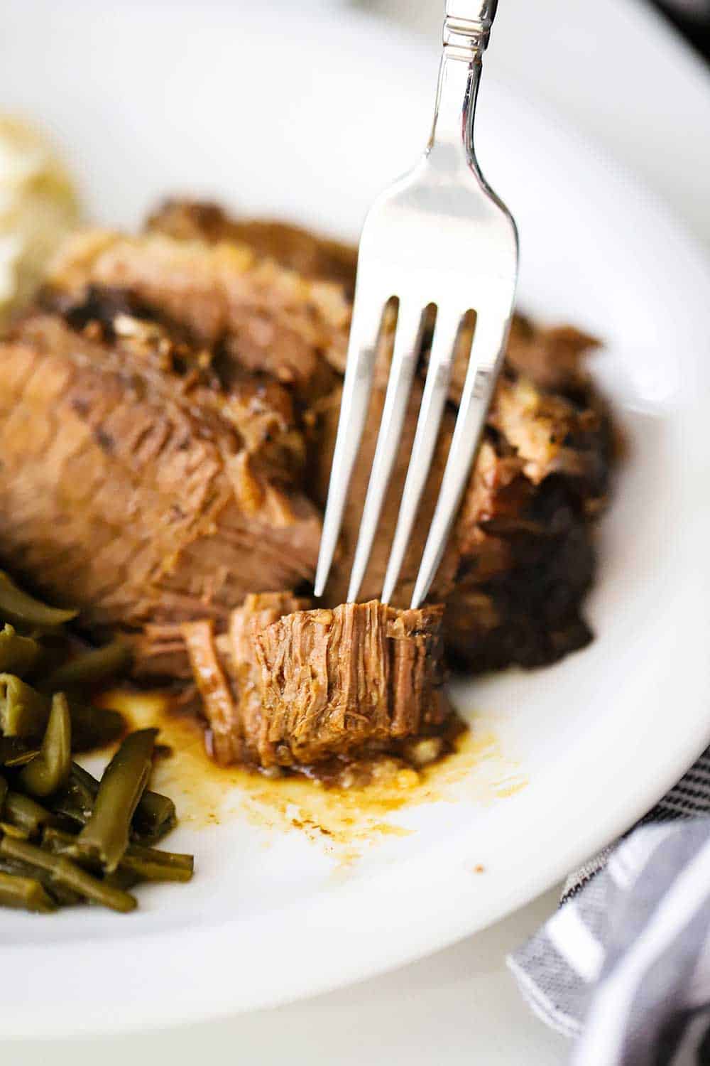 A fork cutting into a tender piece of slow-cooker brisket on a white serving plate. 