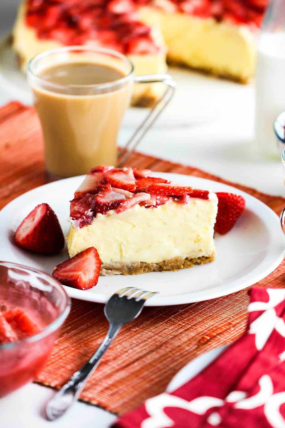 A slice of New York Cheese Cake with Strawberry Glaze sitting next to a cup of coffee and fresh strawberries. 