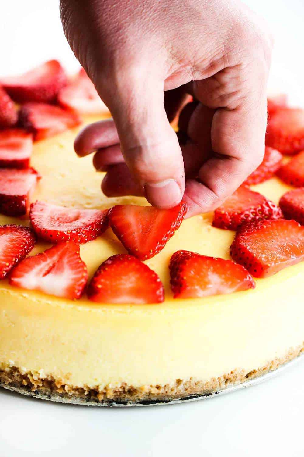 New York Cheese Cake with a hand placing fresh sliced strawberries on top.