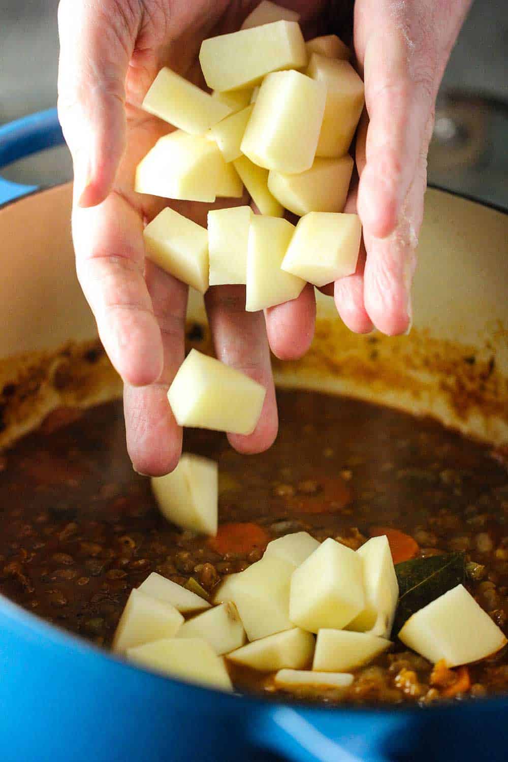 Two hands dropping cubed potatoes into a Dutch oven filled with lentil soup. 