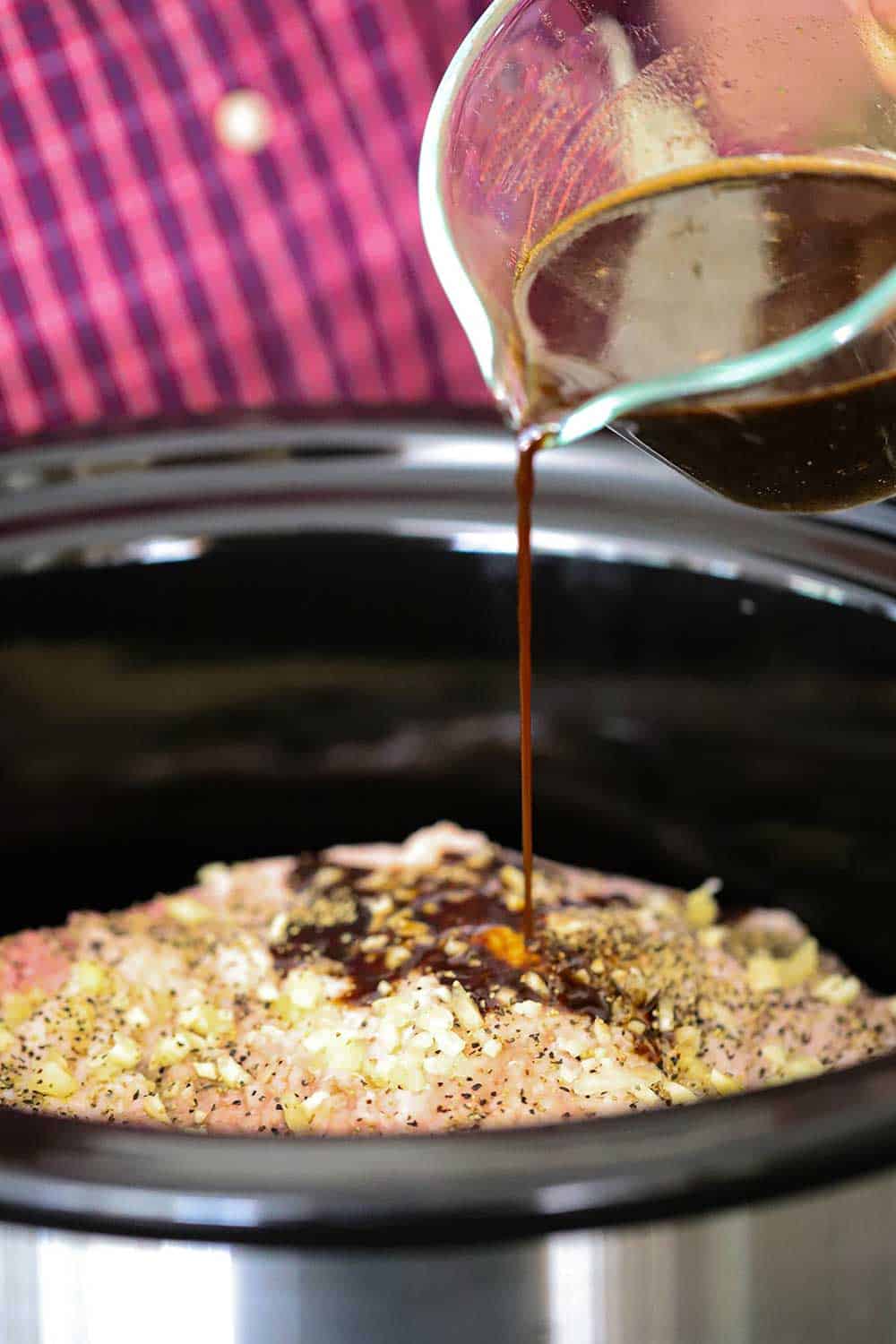 A person pouring a dark marinade over a seasoned brisket in a slow-cooker. 