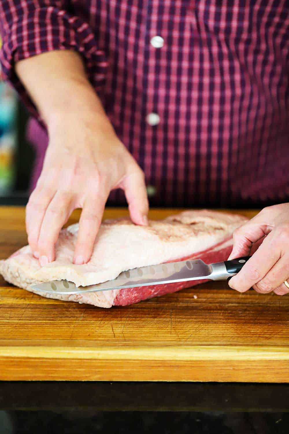 A person using a chef's knife to trim away excess fat on an uncooked brisket on a cutting board. 