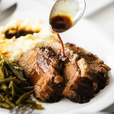 A spoon drizzling au jus sauce on slices of slow cooker brisket on a white plate.