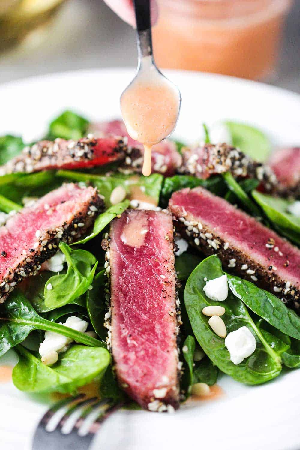 A spoon pouring strawberry vinaigrette over strips of seared tuna steaks on top of a spinach salad on a white plate. 