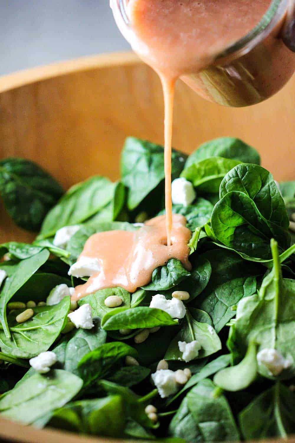 A jar of strawberry vinaigrette being poured into a wooden bowl filled with baby spinach.