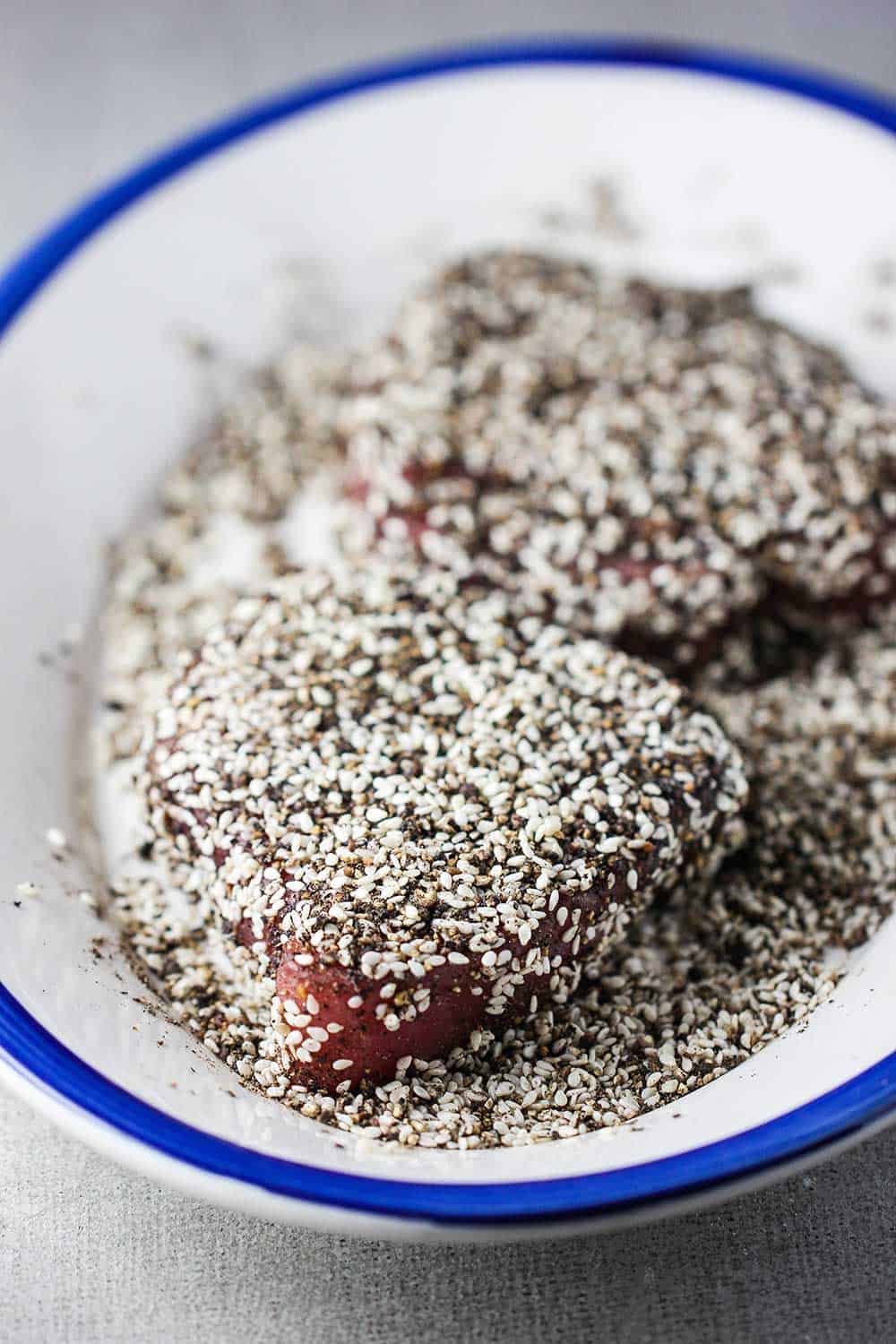 Two raw tuna steaks dredged in ground peppercorns and white sesame seeds on a platter. 