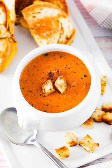 A white soup filled with roasted tomato basil soup next to sliced grilled cheese sandwiches.