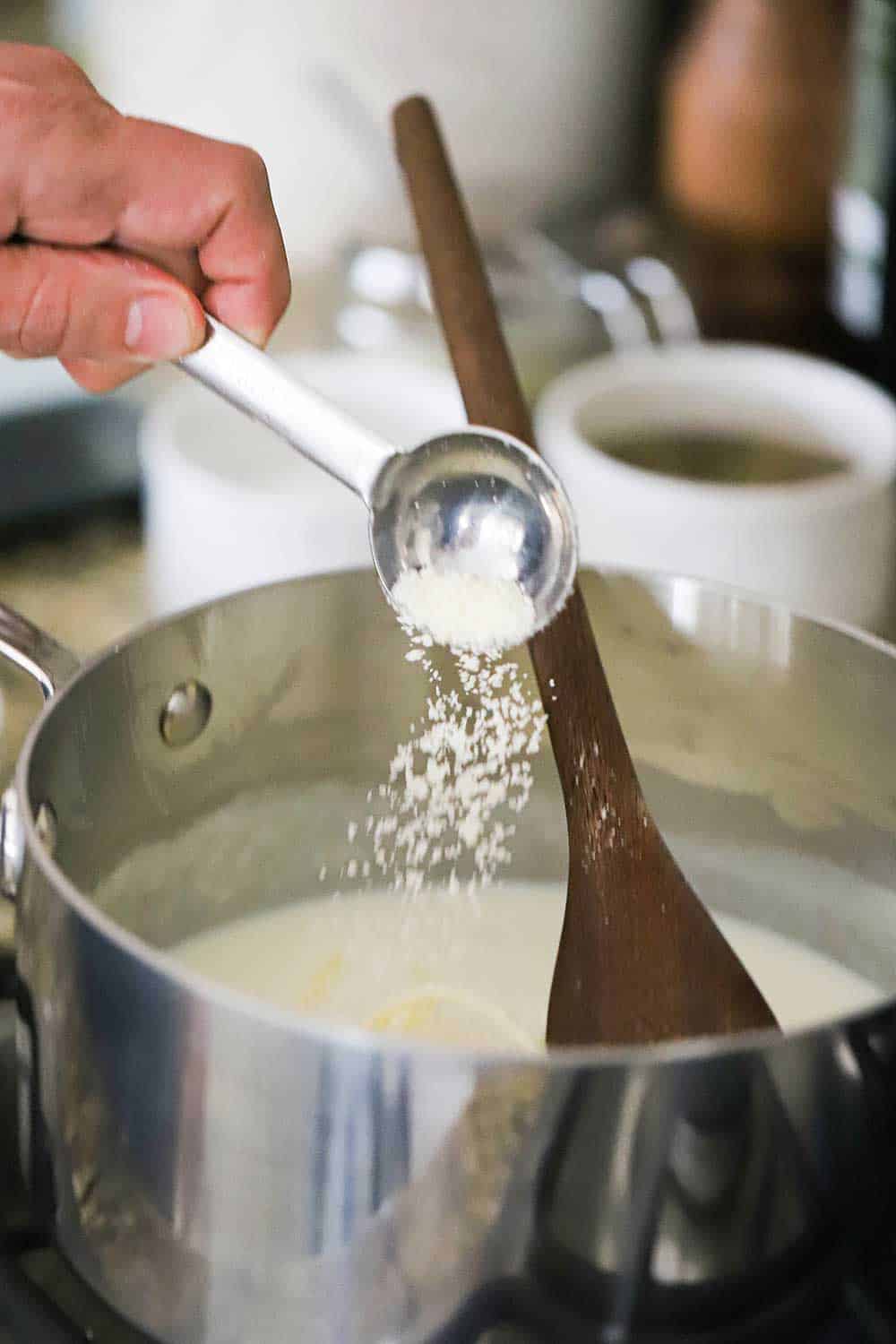 A person sprinkling grated Parmesan cheese from a tablespoon into a saucepan filled with Mornay sauce. 