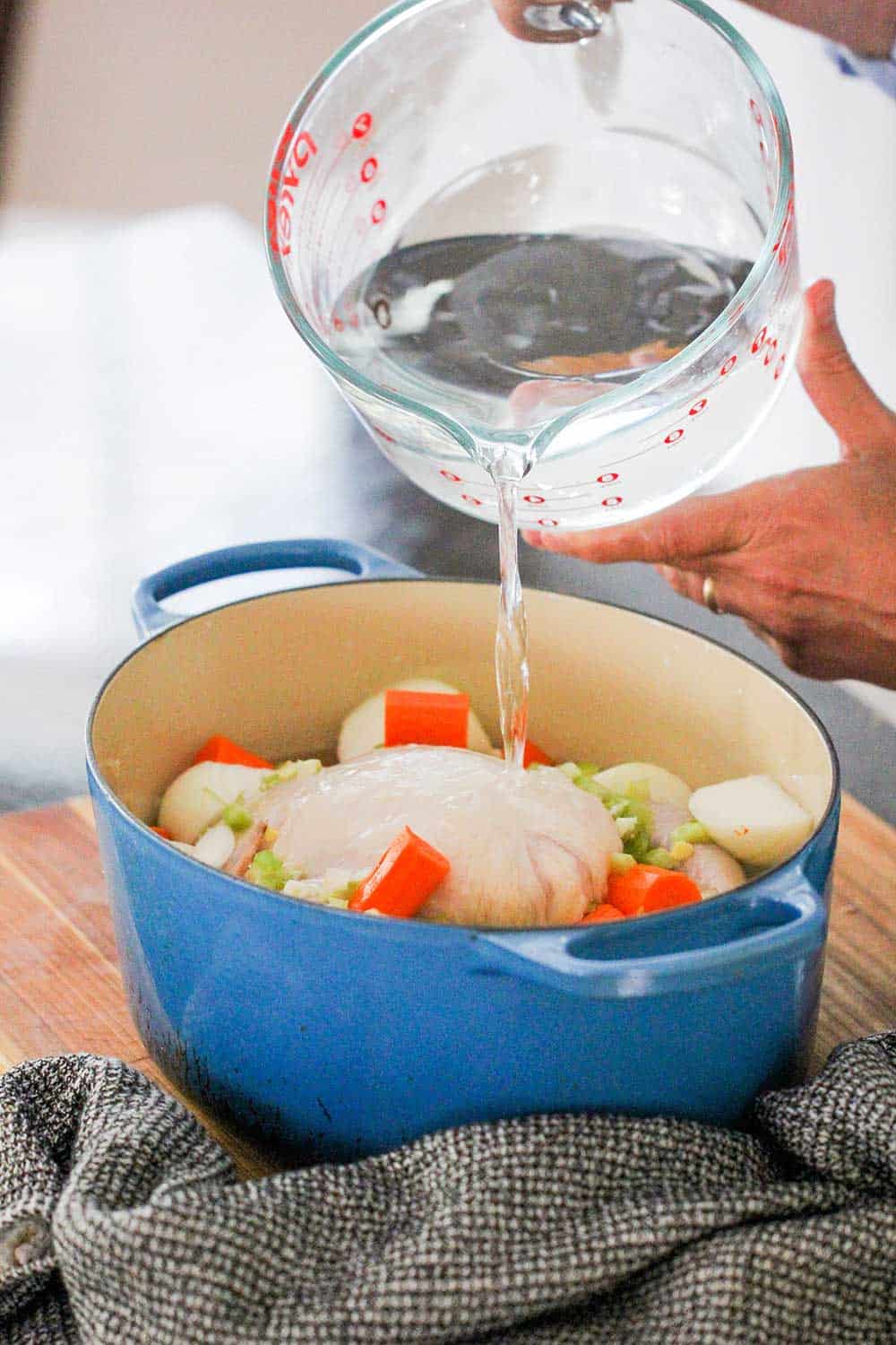 A person pouring a large vessel of water over a chicken and vegetables in a large blue Dutch oven. 