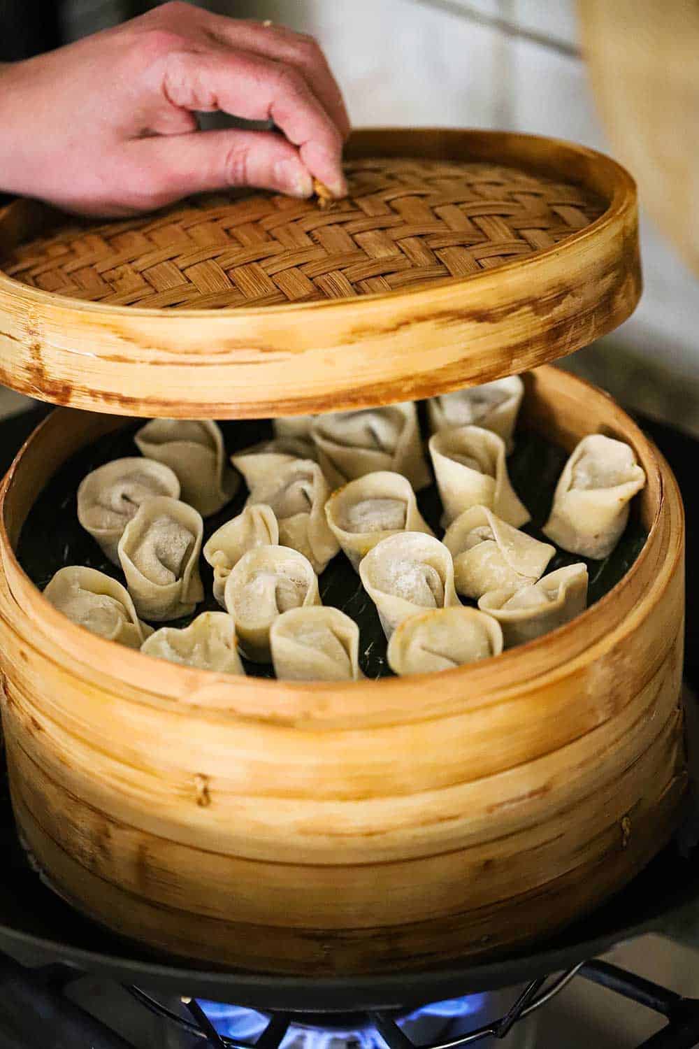 A person placing a bamboo lid onto a bamboo steamer that is filled with vegetarian dumplings all sitting in a wok on a stovetop. 