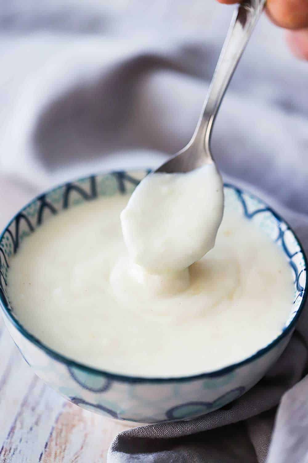 A small serving bowl with a blue pattern on it filled with mornay sauce with a spoon being lifted out of it. 