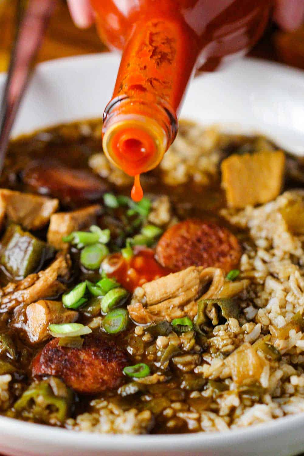A bottle of cayenne hot sauce being poured into a white bowl of chicken and sausage gumbo. 