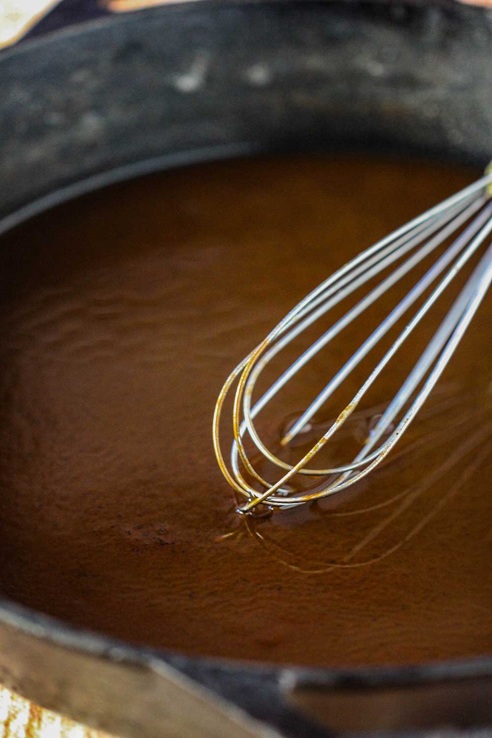 A whisk in a black cast iron skillet containing a dark brown roux