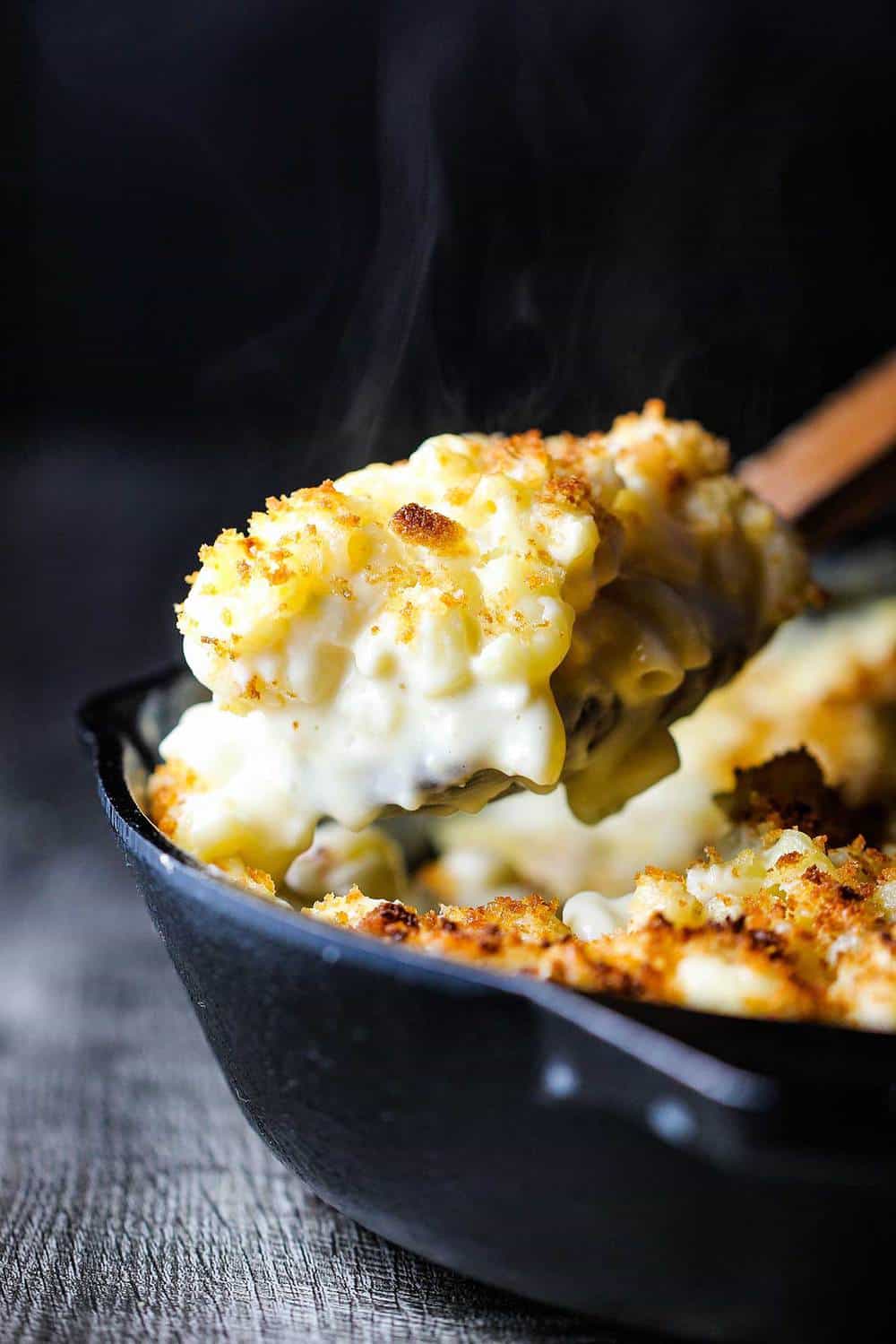 A wooden spoon lifting a helping of ultimate macaroni and cheese from a black cast iron skillet