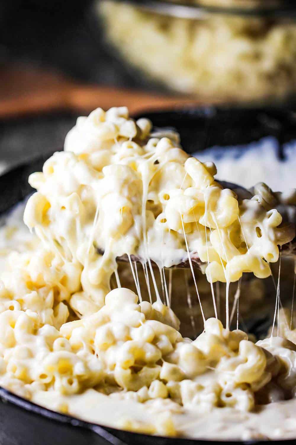 A spoon lifting stringy creamy cheesy mac 'n cheese from a skillet. 