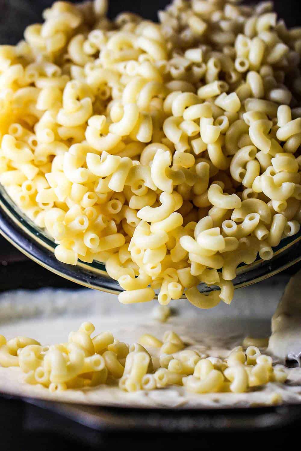 Cooked elbow pasta pouring into a pan of cheesy béchamel sauce. 