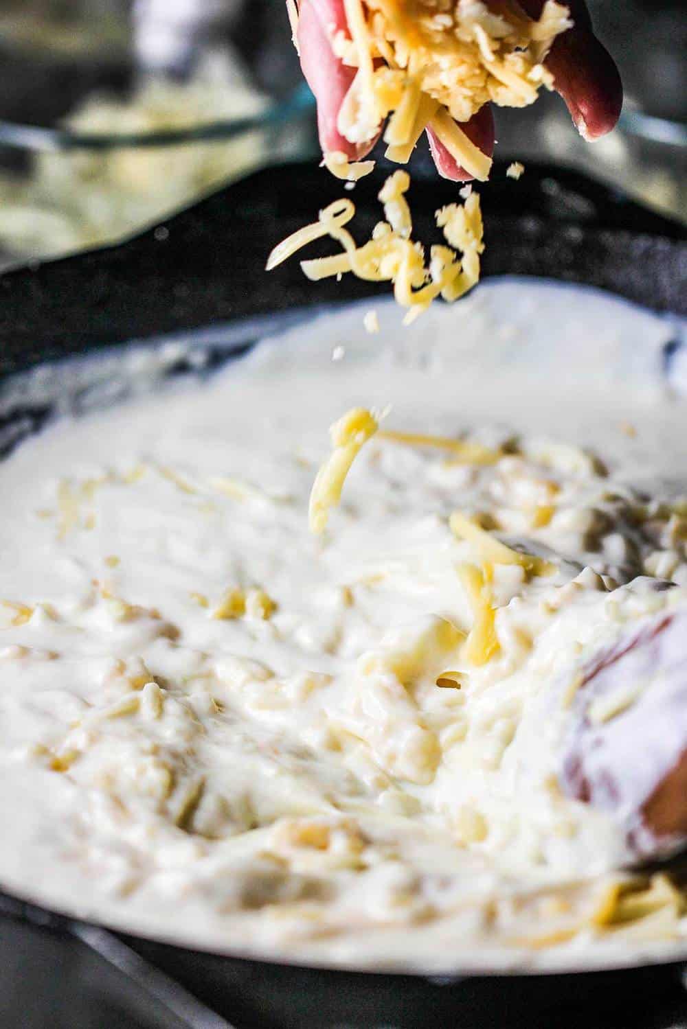 A hand sprinkling in cheese to a béchamel sauce in a cast iron skillet.