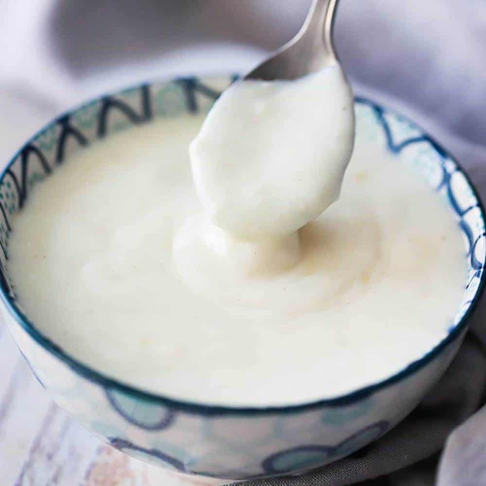 A small serving bowl with a blue pattern on it filled with mornay sauce with a spoon being lifted out of it.