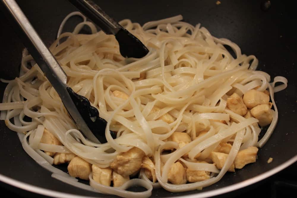 A large black wok filled with cooked chicken pieces and wide Thai noodles with a pair of tongs tossing them. 