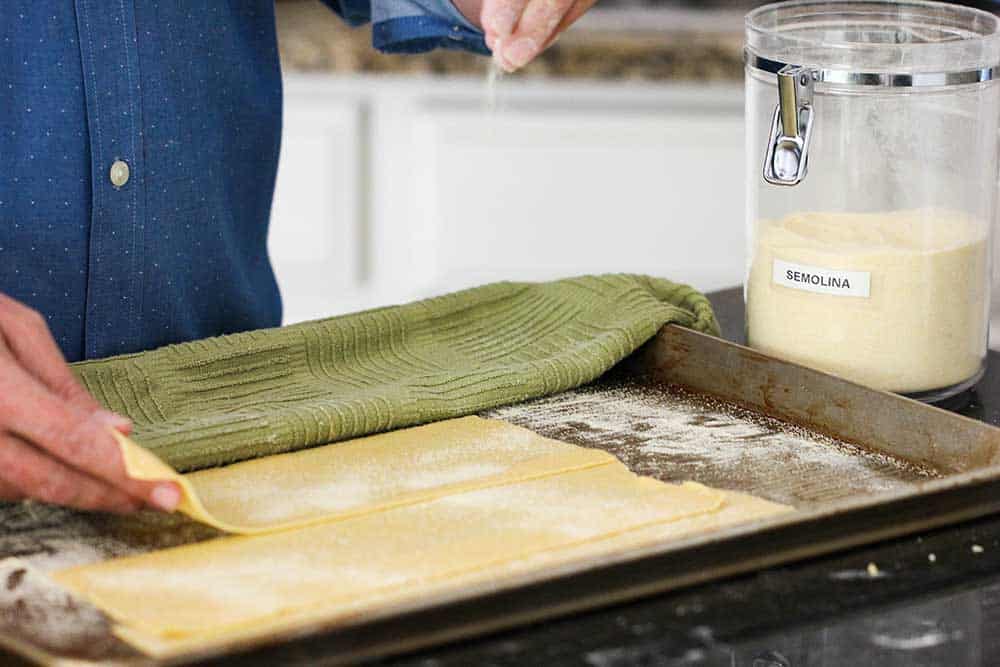 Two hands placing pasta sheets on a baking sheet sprinkled with semolina flour. 