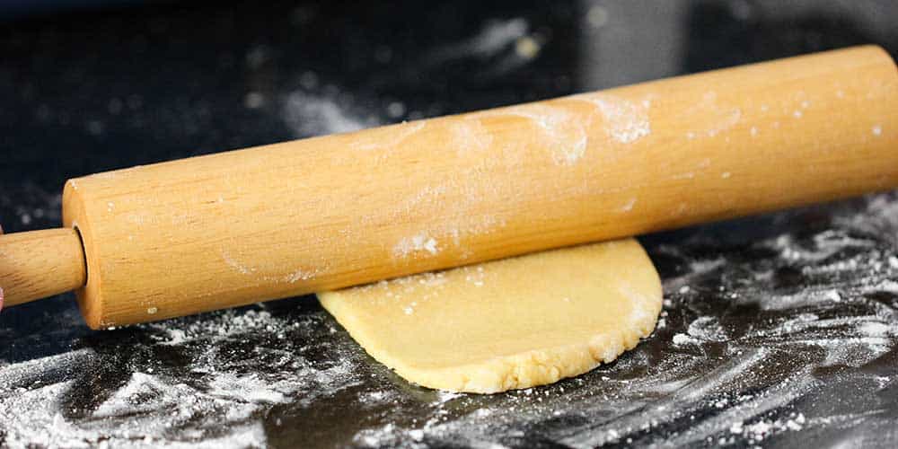 A rolling pin rolling out a piece of homemade pasta dough on a floured workspace. 