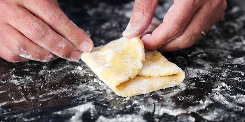 Two hands folding in the sides of a piece of pasta dough on a floured workspace. 